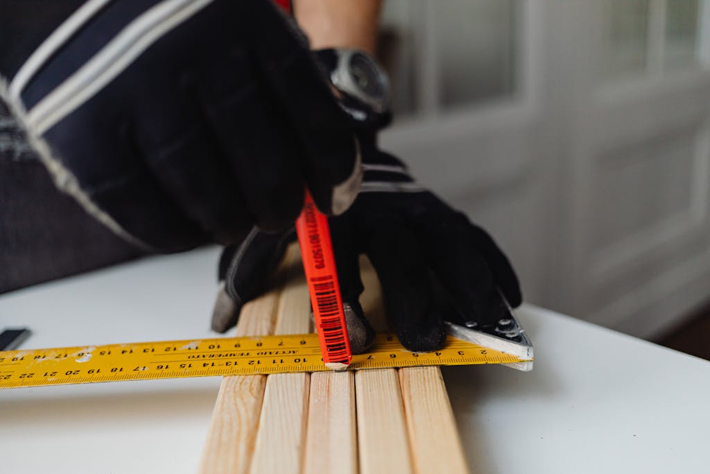 Close-up of a gloved carpenter measuring wood indoors using a ruler and pencil.